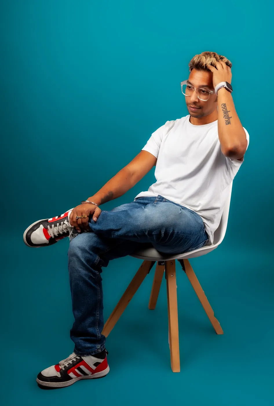Man in a white t-shirt and jeans sitting on a chair against a teal backdrop